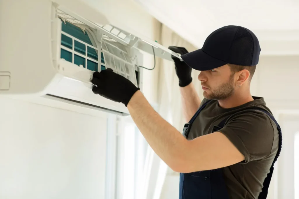 A technician in a navy cap and gloves performs maintenance or replacement on an indoor wall-mounted AC unit.