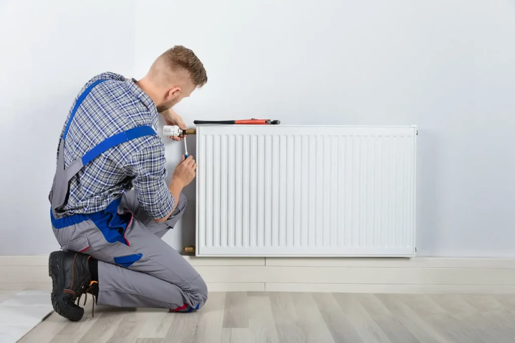 A male technician in gray overalls kneels on a wooden floor, using a screwdriver to service a white radiator mounted on a wall.